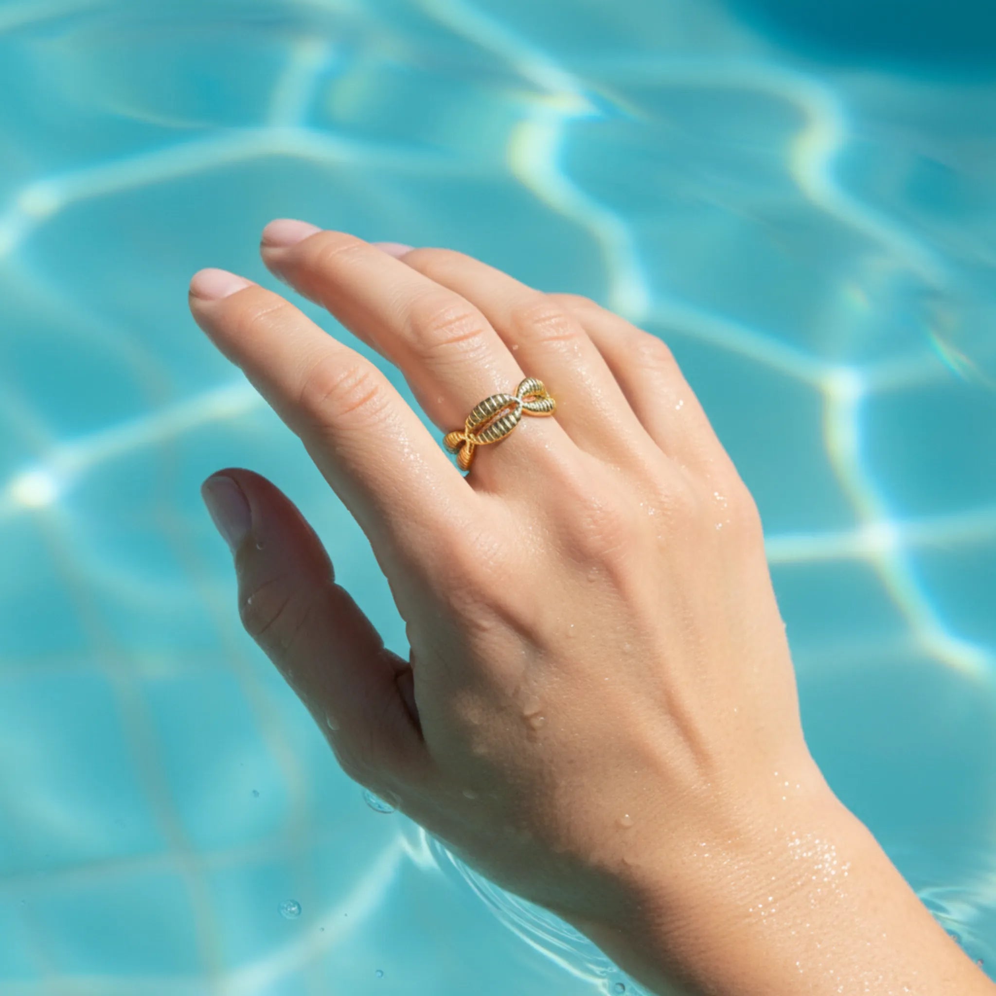 A hand wearing a gold cowrie shell ring in a swimming pool
