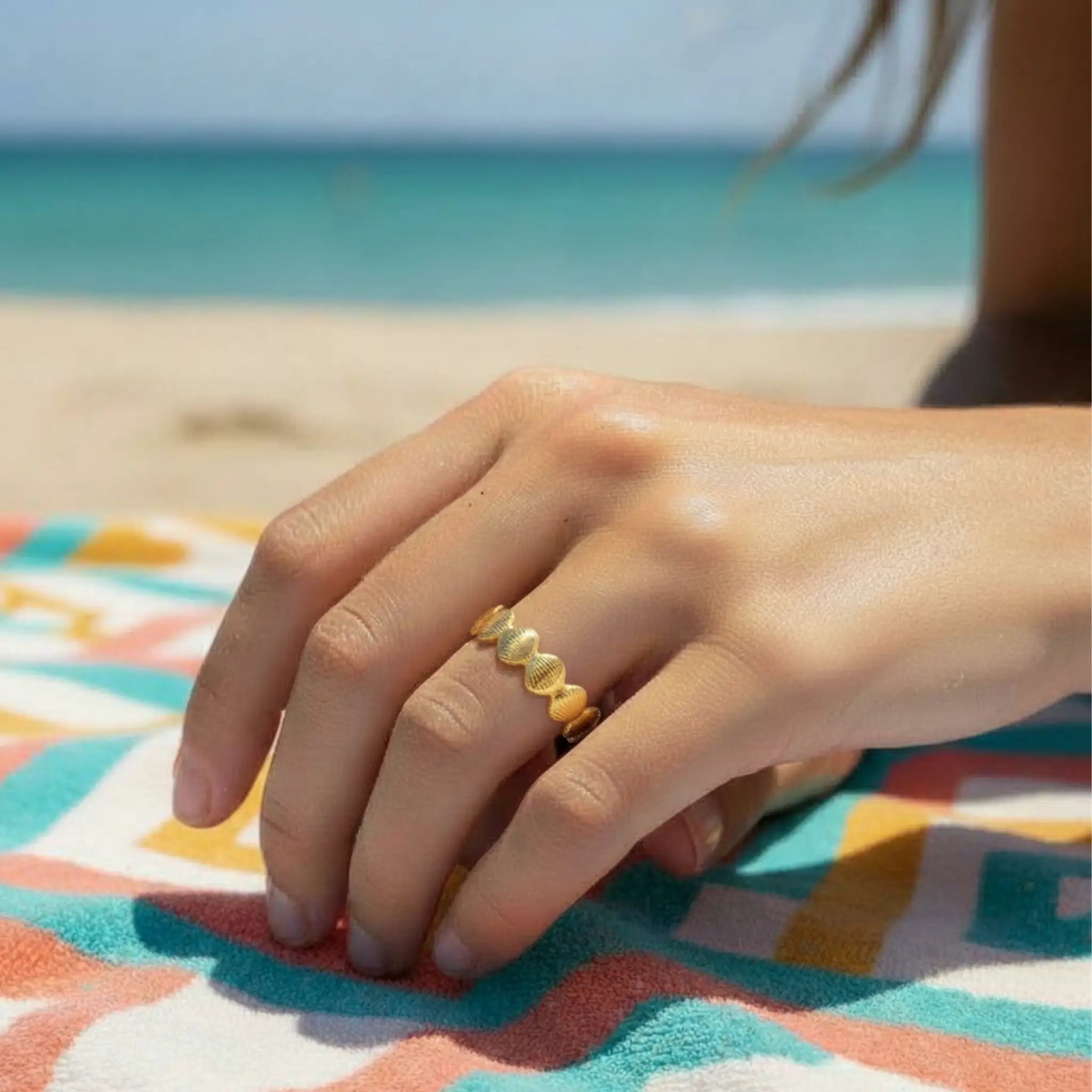 Hand wearing a gold shell band ring on a colorful towel with a beach background