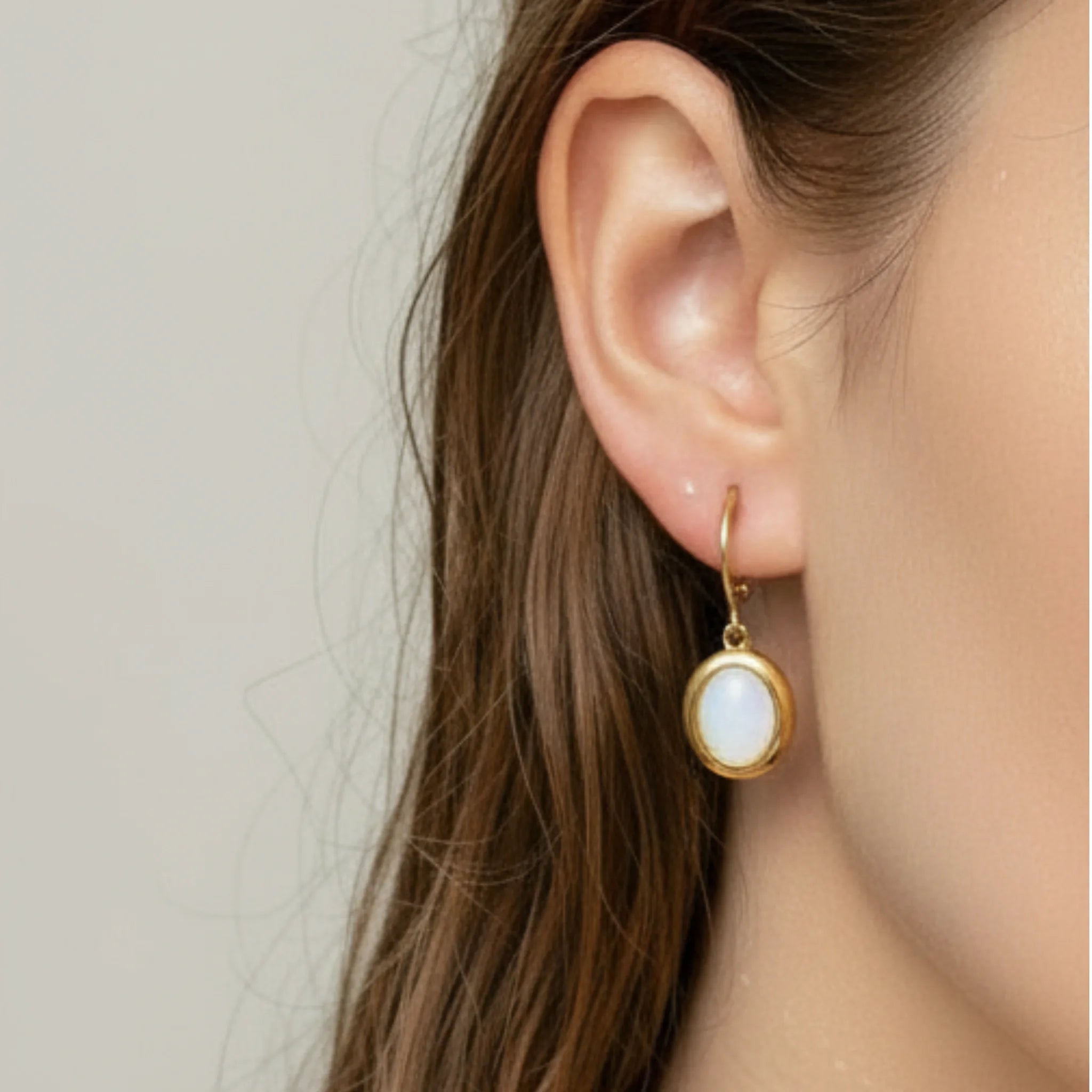 Gold earrings with a white stone worn by a person with long brown hair.