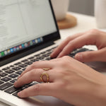 Close-up of hands typing on a laptop with a gold open front sculptural ring on a blurred background