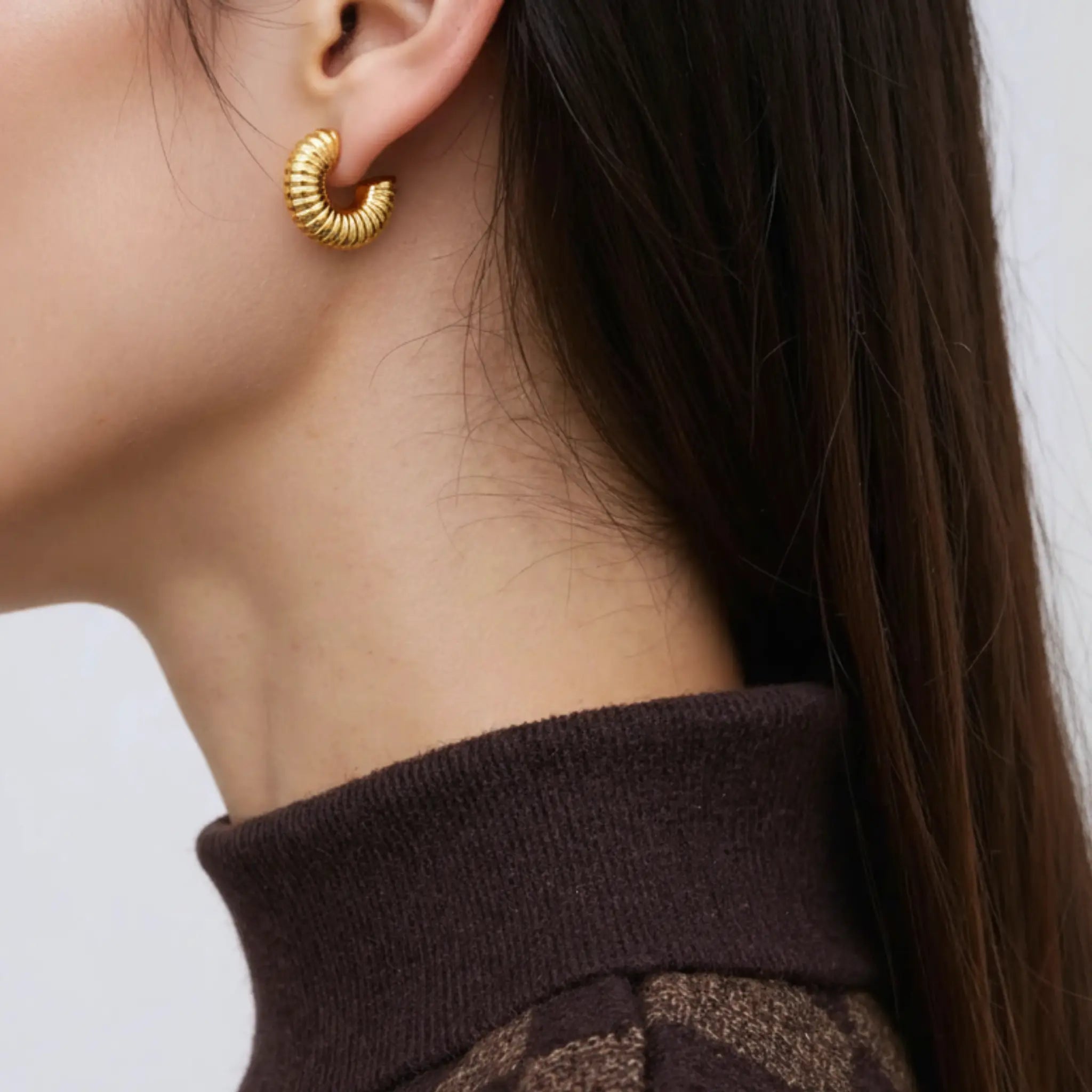Close-up of a person wearing a gold textured spiral hoop earring with a neutral background